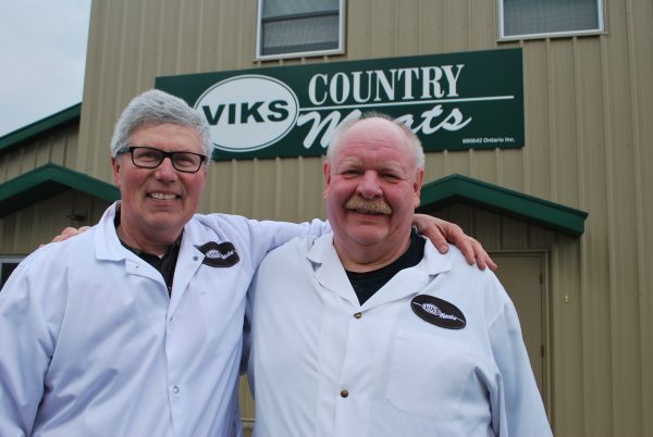 Two men stand infront of a sign for Vik's Country Meat.