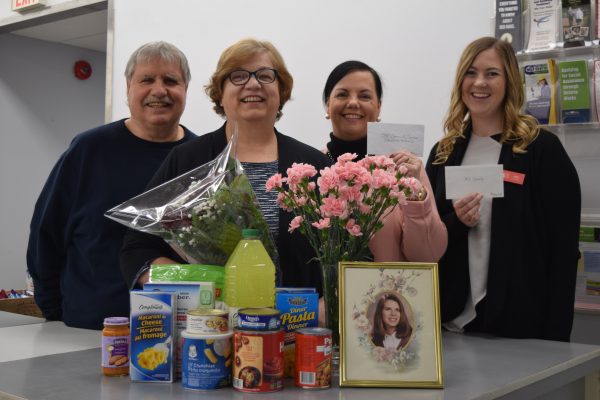 Four people stand behind a collection of flowers, donated food and a framed photo of Christina.