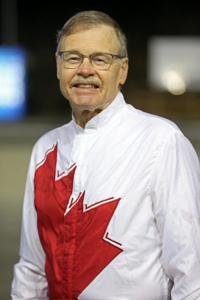 A man wearing glasses and a white riding jacket with a red maple leaf on it smiles at the camera.