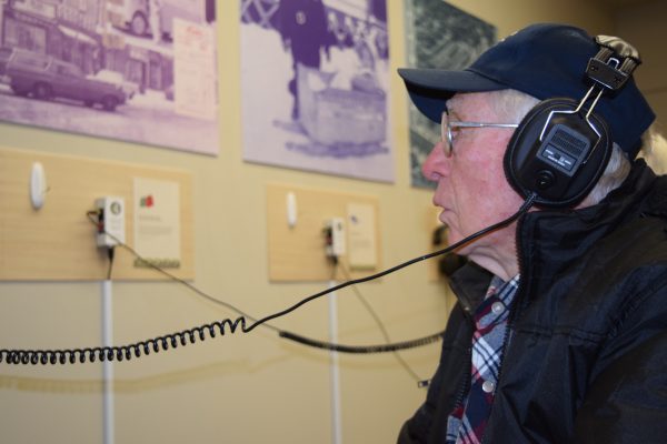 An older man wears headphones while reading text for a museum exhibit that the headphones are attached to.