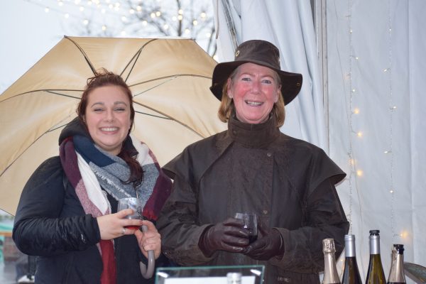 Two women stand under a tent holding on to wine glasses, the one on the left also holding an umbrella.