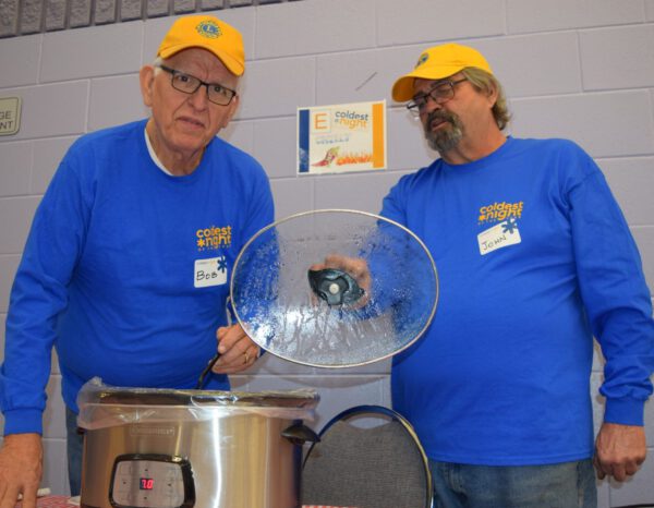 Two men in Lions Club uniforms stand next to a chili pot.