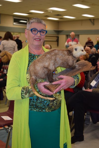 A middle-age woman with close-cropped white hair, blue rim glasses and a green outfit smile while holding a stuffed opossum.