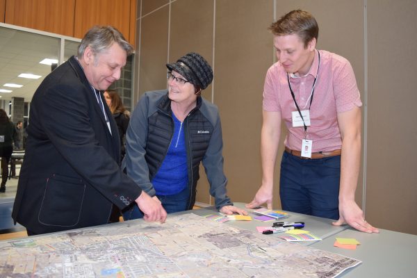 Three people stand around a large map of Smithville, which sits on a table. The man to the left point at the map.