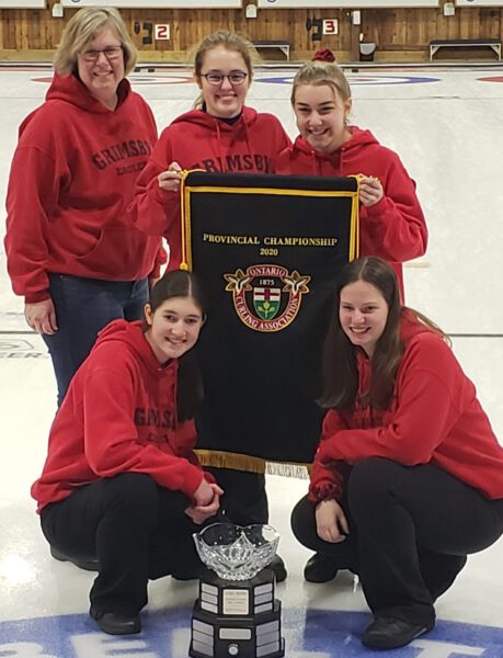 Four girls and their curling coach stand with their champions pendant and trophy.