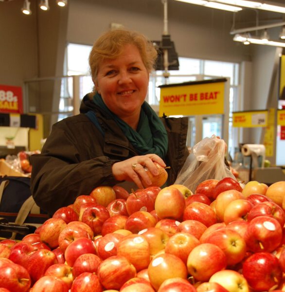 A woman smiles from accross a grocery aisle of apples.