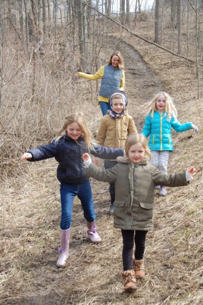 Four young girls and a woman hike down a forested path.