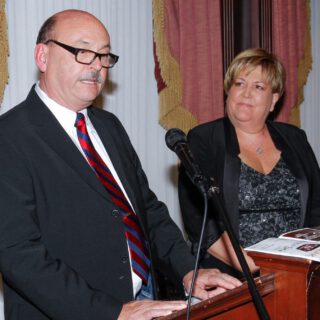 A man speaks at a podium while a woman looks at him from the right.