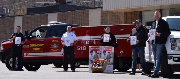 Five individuals stand 2 metres apart in front of a fire truck and a food drive box bearing the logo of the Grimsby Benevolent Fund.