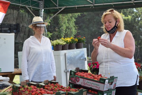A woman watches while another eats a strawberry under a fruit stand tent.